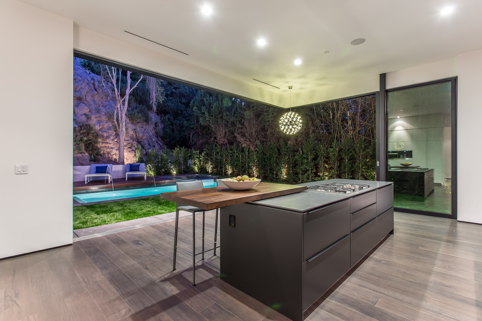 Modern kitchen interior overlooking night-lit garden with pool.