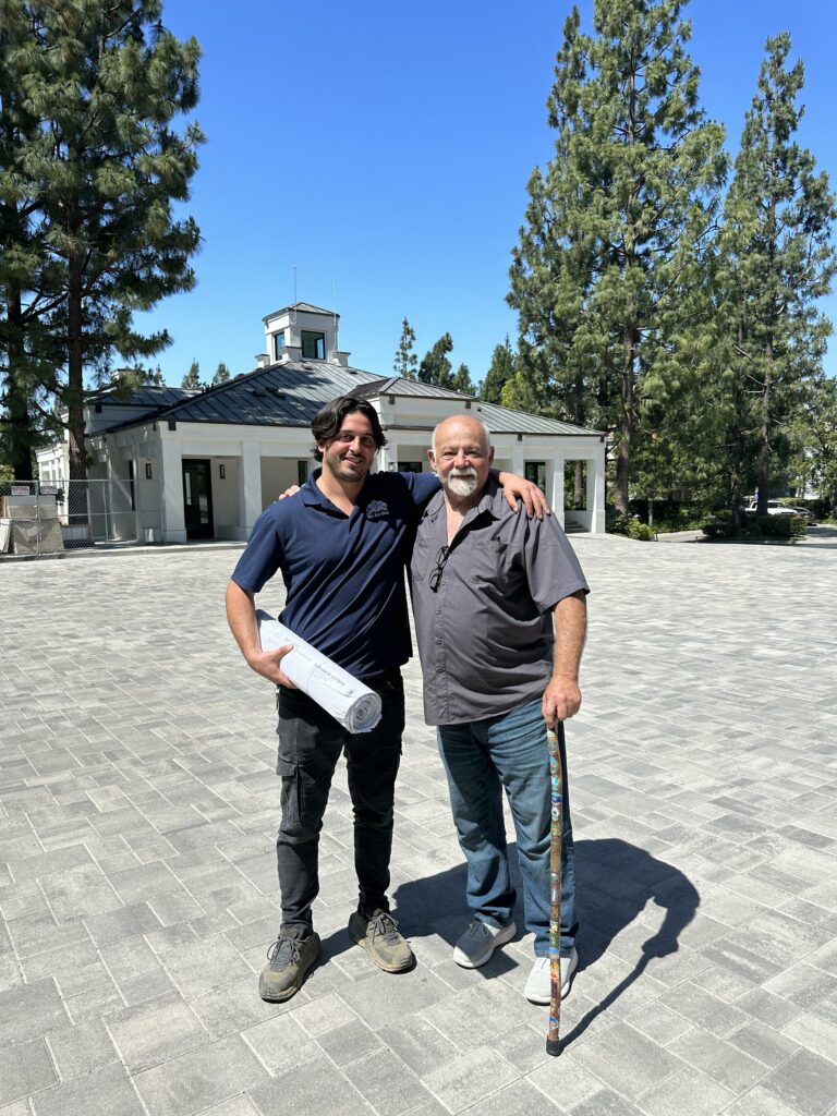 Two men smiling outside near building and trees.