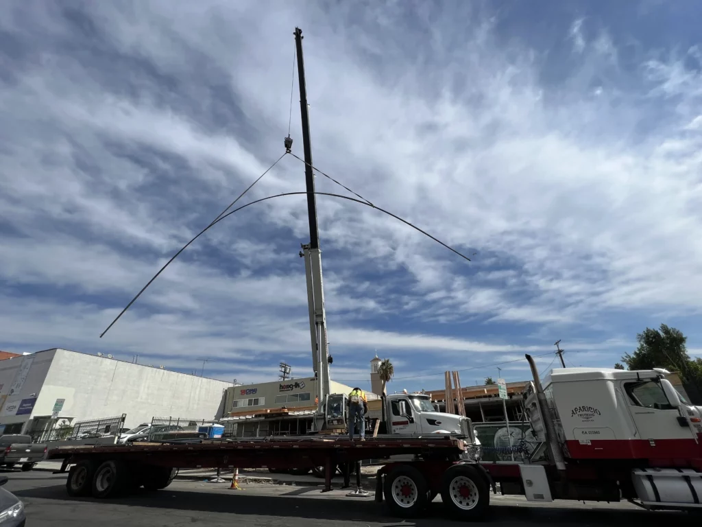 Crane lifting large curved pole onto truck in city.